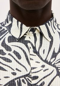 Κοντινό πλάνο πουκάμισου με μαύρο και άσπρο μοτίβο, κουμπωμένο, με αφηρημένα σχέδια φύλλων στο λαιμό και το πάνω μέρος του στήθους ενός ατόμου.