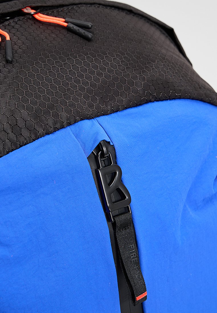 Rucksack mit blauem Stoffkörper, schwarzem sechseckigem Oberpanel und einem verstellbaren schwarzen Riemen mit einer markanten Schnalle und roten Akzenten.