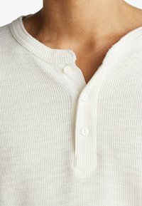 White ribbed henley shirt featuring three buttons at the collar, textured fabric, and a rounded neckline with a slightly irregular edge.