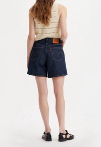 Dark blue denim shorts with a high waist, straight cut, and classic back pockets. Textured, woven beige tank top. Black sandals.