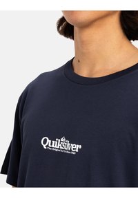 Camiseta de algodón azul marino con cuello redondo, con un logo blanco de Quiksilver en el pecho y mangas cortas. Textura suave, ajuste casual.