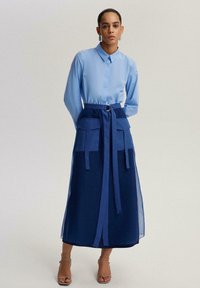 Heleroheline nööbitav särk koos tumesinise pika seelikuga, millel on suured taskud ja vöö, mis on seotud, rõhutades stiilset kihilist disaini.