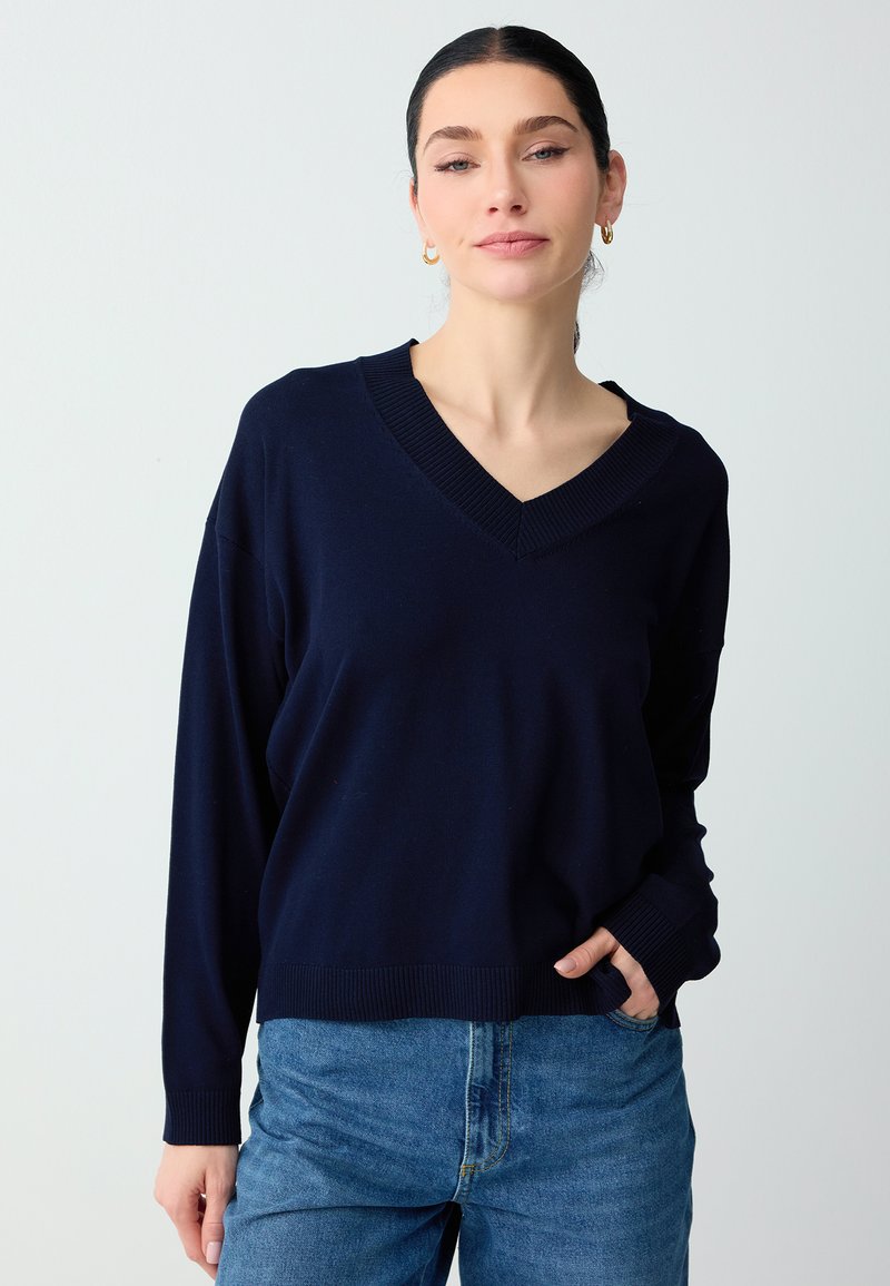 Femme aux cheveux foncés portant un pull bleu marine à col en V et un jean bleu, debout, une main dans la poche, devant un fond clair uni.