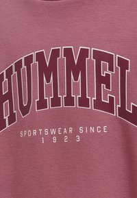 Maroon T-shirt med djärv vit text, "HUMMEL," som bågar över "SPORTSWEAR SINCE 1923," på ett mjukt, strukturerat tyg.