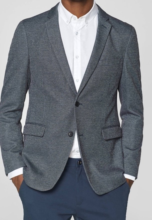 Blazer grigio con texture, rever a pistol, due tasche frontali, chiusura a bottone singolo, indossato sopra una camicia bianca, con pantaloni blu sotto.
