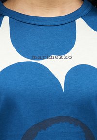 Μπλε βαμβακερό μπλουζάκι με μεγάλο λευκό floral σχέδιο και λογότυπο "marimekko", με στρογγυλή λαιμόκοψη και κοντά μανίκια.