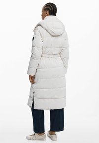 Lange pufferjas in lichtbeige met een capuchon, elastische taille en drukknoopsluiting aan de zijkanten. Gemaakt van een glad, gewatteerd materiaal.