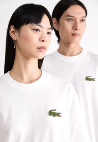 Tricourile din bumbac alb au un mic logo verde cu un crocodil pe piept. Designul include un guler clasic rotund și mâneci scurte.
