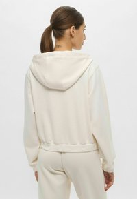 Felpa cropped in morbido tessuto crema, con cappuccio, polsini elastici e vita elasticizzata. Texture liscia con cuciture minime per un design pulito.