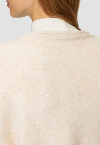 Beiger, gestrickter Pullover mit weicher Textur, ausgestattet mit einem entspannten Kragen und einem subtilen Melange-Muster. Nahtdetail am Ausschnitt.