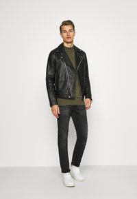 Chaqueta de cuero negro de motorista con cremalleras, suéter verde oliva, jeans oscuros y zapatillas blancas. El modelo está de pie contra un fondo blanco liso.