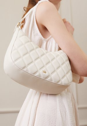 Gestepptes cremefarbenes Lederhandtasche mit geschwungener Form, goldfarbenen Beschlägen und verstellbarem Riemen, gehalten vor einem hellen, strukturierten Stoffhintergrund.