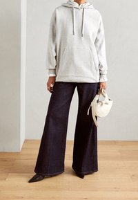 Sudadera con capucha de color gris claro con un bolsillo tipo canguro, combinada con pantalones vaqueros oscuros de pierna ancha. Sosteniendo una pequeña bolsa blanca, con zapatos negros planos.