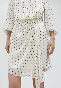 Vestido blanco con lunares negros, con mangas tres cuartos, cintura ceñida con cinturón, y una textura fluida y transparente.