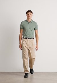 Ung mann går innendørs iført en grønn polo skjorte, beige bukser, svart belte, og svarte joggesko mot en hvit bakgrunn.