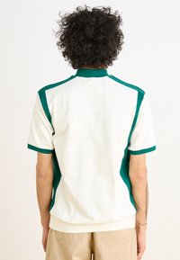 Πολυκόκκινη μπλούζα πόλο κοντό μανίκι σε κρεμ απόχρωση με πράσινες λεπτομέρειες στους ώμους και στο γιακά. Φτιαγμένη από μαλακό ύφασμα, διαθέτει άνετη εφαρμογή και ribbed τελείωμα.