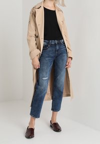 Kvinna i beige trenchcoat, svart topp, korta blå jeans med ett litet hål och mörkbruna loafers, stående mot en enfärgad bakgrund.