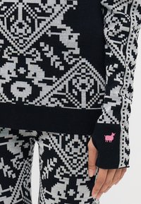 Svart og grå mønstret genser med intrikate designs; inkluderer en rosa saue-logo på mansjetten. Materialet ser mykt og teksturert ut.