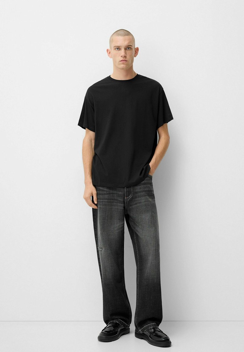 T-shirt en coton noir à manches courtes, associé à un jean noir délavé à coupe ample et des chaussures noires. Design simple, sans motifs visibles.