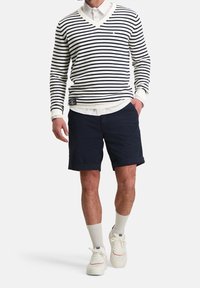 Striped navy and white v-neck sweater layered over a white shirt, paired with navy shorts. White sneakers and light socks complete the look.
