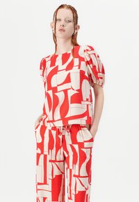 Červený a krémový outfit se topem s nadýchanými rukávy a širokými kalhotami s geometrickým vzorem abstraktních tvarů a linií.