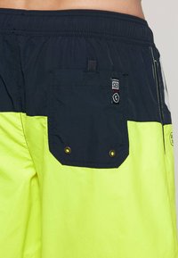 Laivastonsiniset ja kirkkaankeltaiset uimapuvut, joissa on teksturoitu pinta, takatasku, metalliset reiät ja logopatch vyötäröllä.