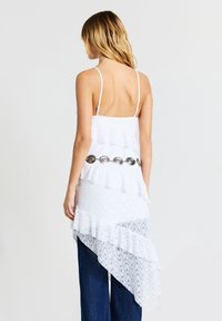 Top bianco in pizzo a strati con spalline sottili, orlo asimmetrico e una cintura decorativa argentata. Il tessuto testurizzato aggiunge interesse visivo.