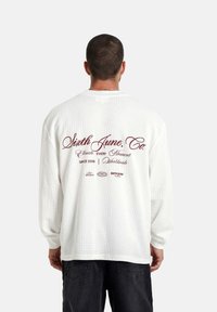 Man met kort haar, gekleed in een witte shirt met lange mouw en bordeauxrode tekst "Sixth June, Co." en zwarte broek, gezien van achteren.