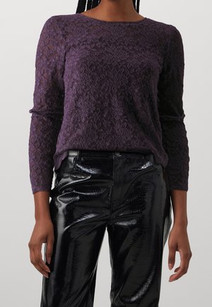 Femme portant un haut en dentelle violet à manches longues et un pantalon noir brillant texturé, debout, les bras détendus le long du corps.