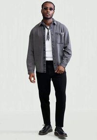 Hombre con gafas de sol negras, camisa de botones gris sobre una camiseta blanca, pantalones negros, zapatos negros y una bufanda de cuello negra con patrón, de pie contra un fondo blanco.