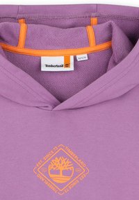 Haut à capuche violet en coton, arborant un logo Timberland orange dans un motif de diamant. Comprend une étiquette orange à l'intérieur du col.