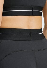 Black athletic top featuring a wide waistband with white stripes. The fabric has a smooth texture and a fitted design with flat seams.