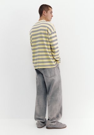 Young man facing sideways wearing a yellow and black striped long-sleeve shirt, loose gray jeans, and gray sneakers against a white background.