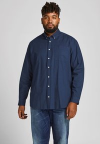 Marineblaue Button-Down-Hem mit spitzem Kragen, Brusttasche und langen Ärmeln; aus strukturiertem Baumwollstoff gefertigt. Kombiniert mit Jeans.