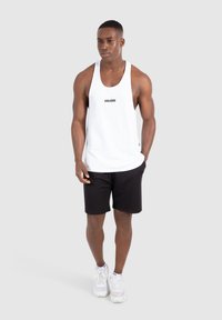 Weißes ärmelloses Tanktop mit schwarzem Text "SMILODOX", kombiniert mit schwarzen Shorts und weißen Sportschuhen. Neutraler Hintergrund.