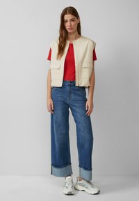 Lichtbeige mouwloos vest met voorzakken, gecombineerd met een rode t-shirt en brede blauwe jeans. Witte sneakers met grijze accenten.