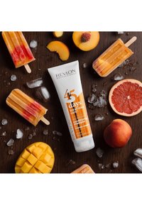 Tube de shampooing revitalisant Revlon Professional, blanche avec des accents oranges, à côté de mangue, tranches de pêche, sucettes glacées fruitées et cubes de glace sur du bois.