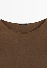 Haut marron en tricot avec un large col rond et une étiquette noire cousue à l'intérieur affichant les détails de la marque et de l'origine.