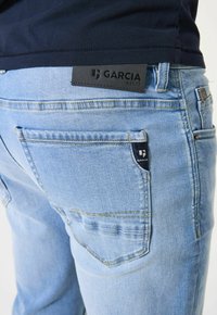 Jeans de mezclilla azul claro con un parche de cuero negro etiquetado como "GARCIA EST.72" en la cintura, con bolsillos traseros visibles y detalles de costura.