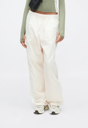 Personne portant un pantalon blanc ample, des baskets blanches et beiges, un haut à manches longues vert olive et une banane noire sur fond uni.