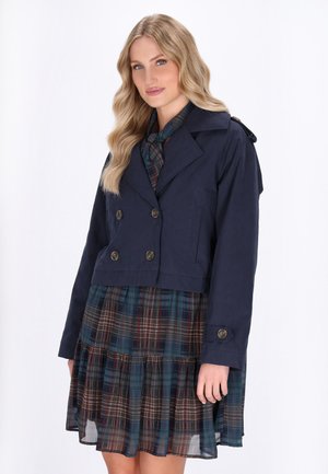 Giacca doppiopetto cropped di colore navy con grandi bottoni, caratterizzata da un colletto a punta e spalline, indossata sopra un vestito a quadri.