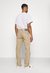 Jeans de mezclilla beige con corte recto, costuras visibles, bolsillos traseros y una etiqueta de marca negra en la cintura. Llevados con una camiseta gris claro.