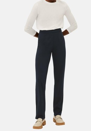 Person iført sorte bukser med høj talje og lige ben, cremefarvet langærmet top og beige loafers med tyk sål, stående med hænderne bag ryggen.