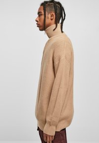 Beige strikkegenser med høy krage, med ribbestrikket tekstur og senkede skuldre. Genseren har en avslappet passform og lange ermer.
