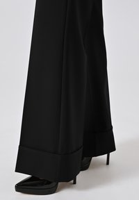 Pantaloni neri a gamba larga realizzati in tessuto morbido, con orlo risvoltato, abbinati a tacchi a punta neri, creando una silhouette elegante.