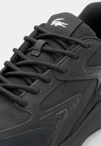 Black athletic shoe with textured synthetic upper, featuring dark laces and a signature logo on the tongue. Close-up of material details.