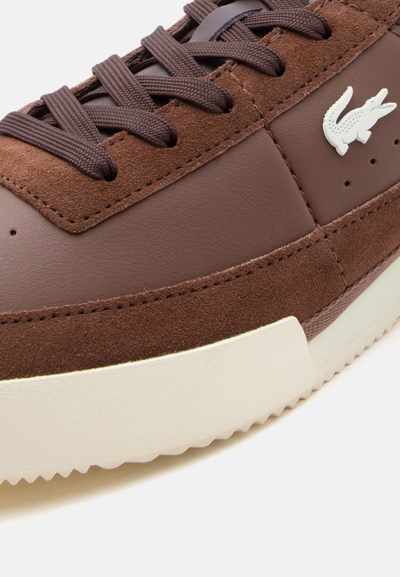 Bruine sneaker met suède en leren panelen, bruine veter, witte gestructureerde zool en wit krokodillenlogo aan de zijkant.