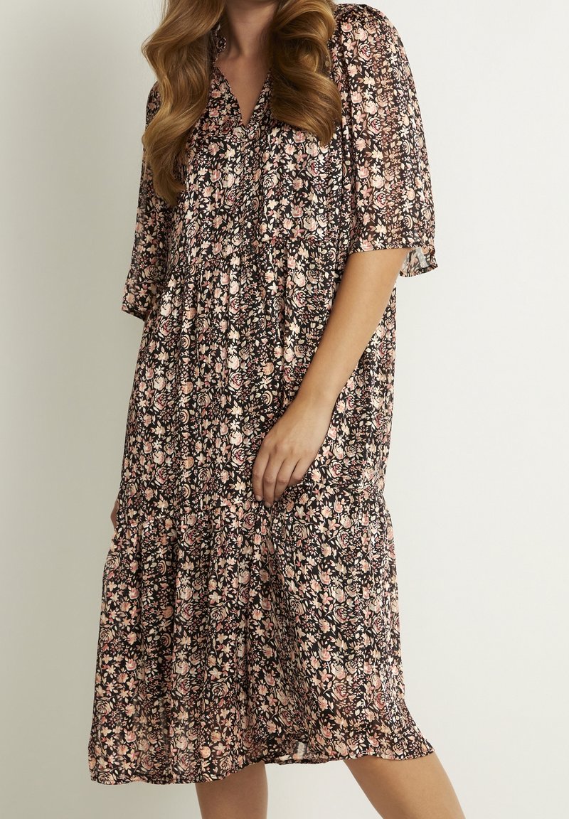 Schwarzes Blumenkleid mit kurzen Ärmeln, das mit rosa und weißen Blumenmustern, einem V-Ausschnitt und einem gestuften Saumdesign versehen ist.