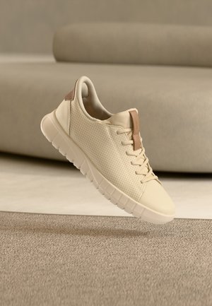 Zapatilla beige con acabado de cuero liso, lados perforados y un acento en tono marrón claro en el talón. Presenta una suela texturizada y diseño con cordones.