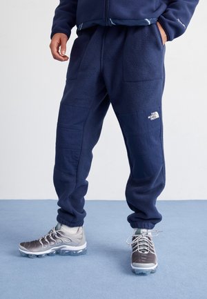 Person iført mørkeblå fleece-joggingbukser med forstærkede knælapper og grå atletiksneakers, der står på blåt tæppe med hænderne i lommerne.